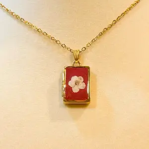 Noelia Rectangle Floral Locket Necklace with Gold Chain and Red Background Featuring a White Flower Design