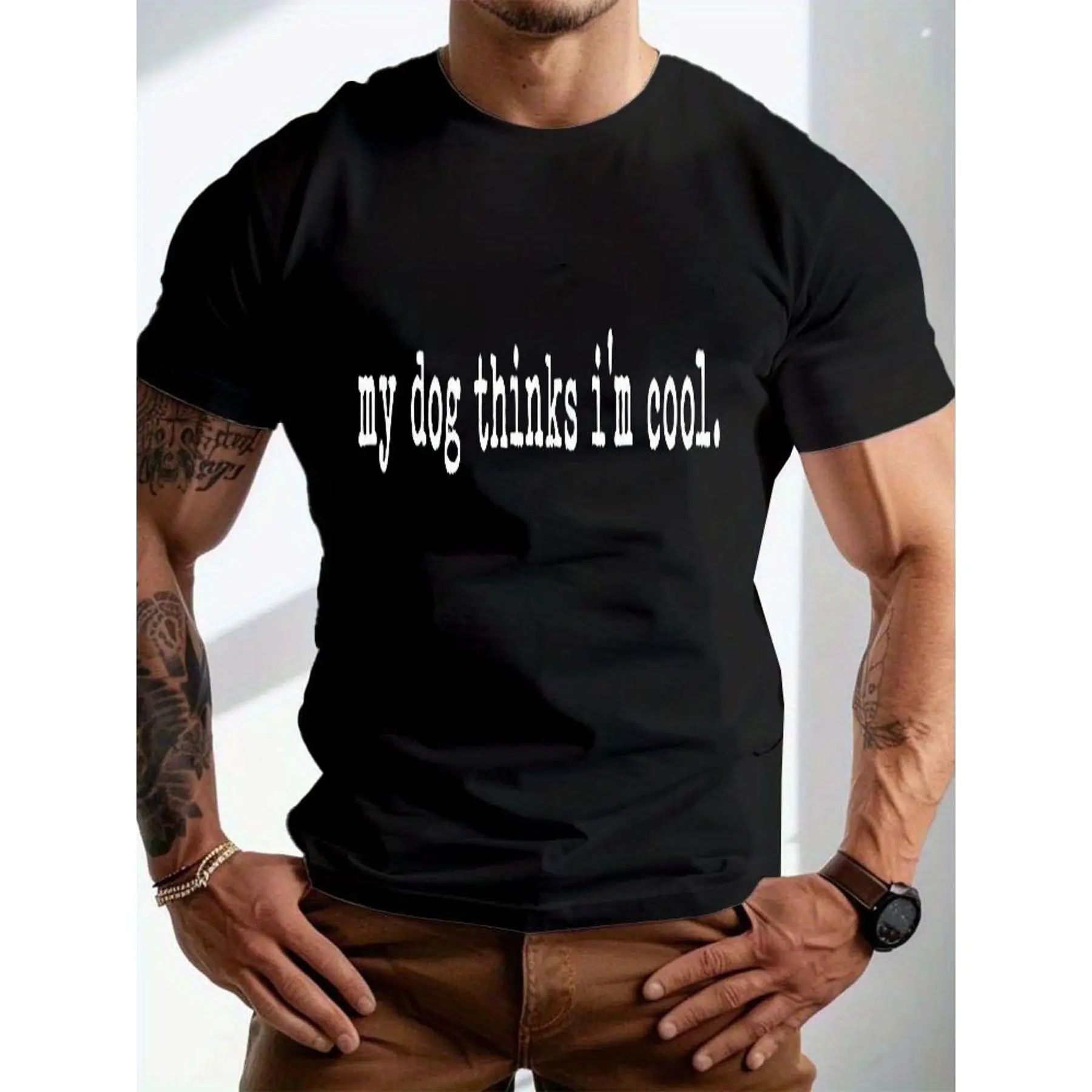 A Muscular Male Model Dressed in a Snug Black Short-Sleeve Crewneck T-Shirt Featuring a Typewriter-Style Print That Says 'My Dog Thinks I'M Cool' on The Chest, Paired with Tan Straight-Leg Trousers, Golden Chain Bracelets, And a Leather Wristwatch