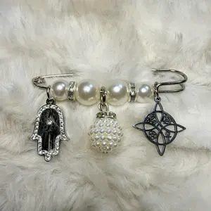 Beautiful silver-tone brooch decorated with soft pearl beads, crystal accents, and meaningful charms. Features a Hamsa hand, pearl sphere pendant, and Celtic-inspired charm for protection, harmony, and spiritual strength.