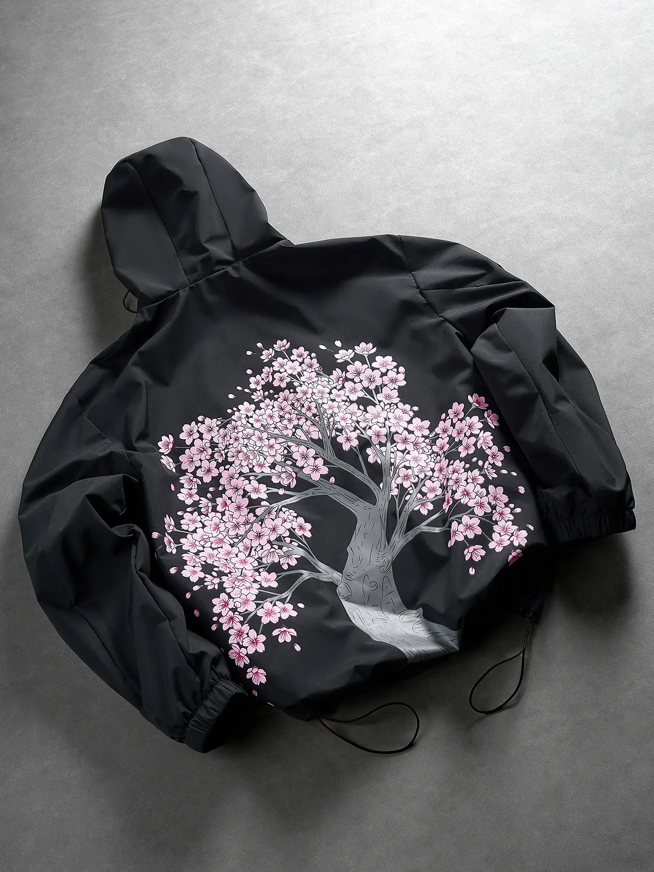 Men’s hooded windbreaker casual jacket featuring an allover print of cherry blossom trees across the back; a black base with pink flowers creates a striking visual contrast that breaks away from the somber feel of a plain black piece. It delivers a strong