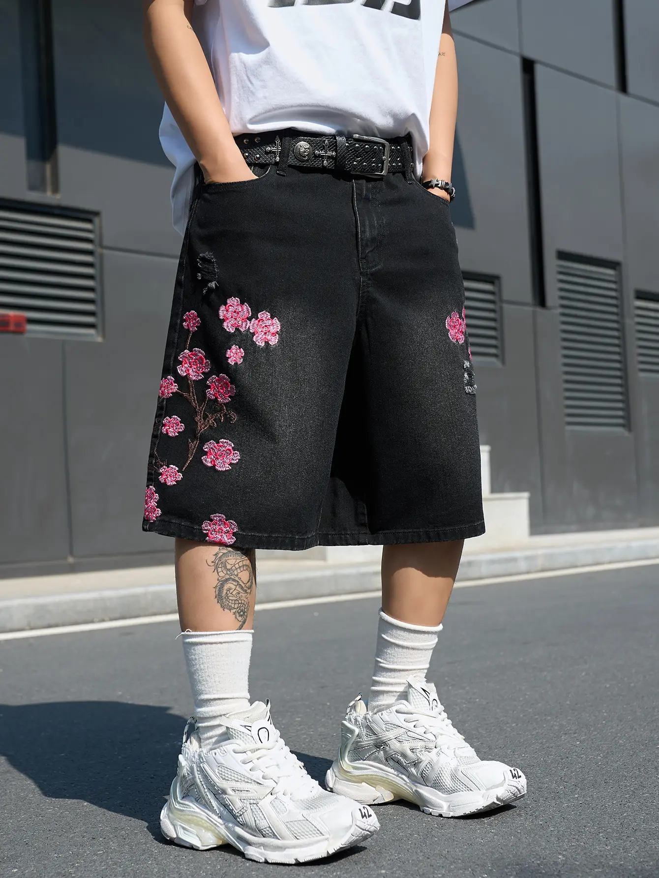 Men’s black denim shorts with pink cherry blossom embroidery and a distressed look, featuring a loose wide-leg fit, mid-rise waist, floral print, and knee length. Durable, multi-pocket streetwear bottoms in Harajuku style, suitable for everyday wear durin