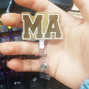 A work card telescopic buckle with "MA" letters, the main body is based on golden glitter as the background, equipped with a telescopic structure and a transparent clip, which can be used to hang work cards, suitable for scenes that need to display signs.