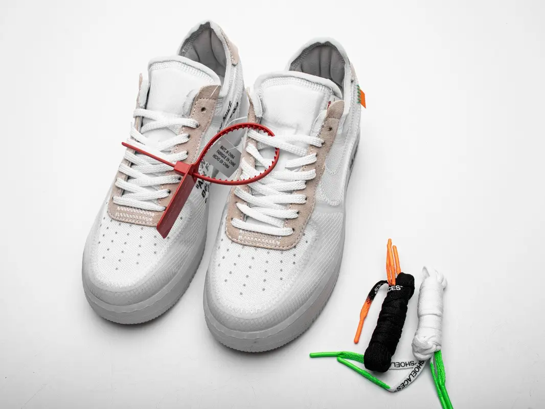 Off‑white low‑top casual sneakers with beige panel stitching details, red plastic lock fastener on laces, and spare colorful shoelace accessories placed beside the footwear
