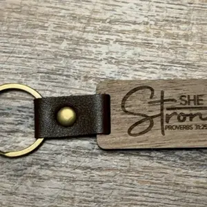 “She is Strong” Wooden Keychain with brass colored keyring attached with brown leather. Measures 1”x 2”. Single sided keychain. Daily Daily