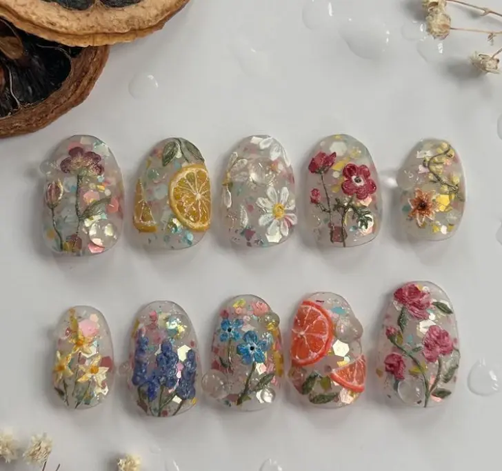 Fresh botanical nails with pressed flowers, citrus slices, and iridescent flakes encapsulated in clear gel for a bright, garden-inspired spring aesthetic