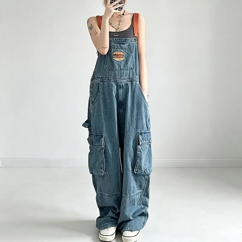 Y2K vintage workwear-style women’s denim bib overalls with a splatter-paint dot design, leather chest patch, large-capacity cargo pockets on both sides, adjustable shoulder straps, loose wide fit, hip-hop everyday trend.