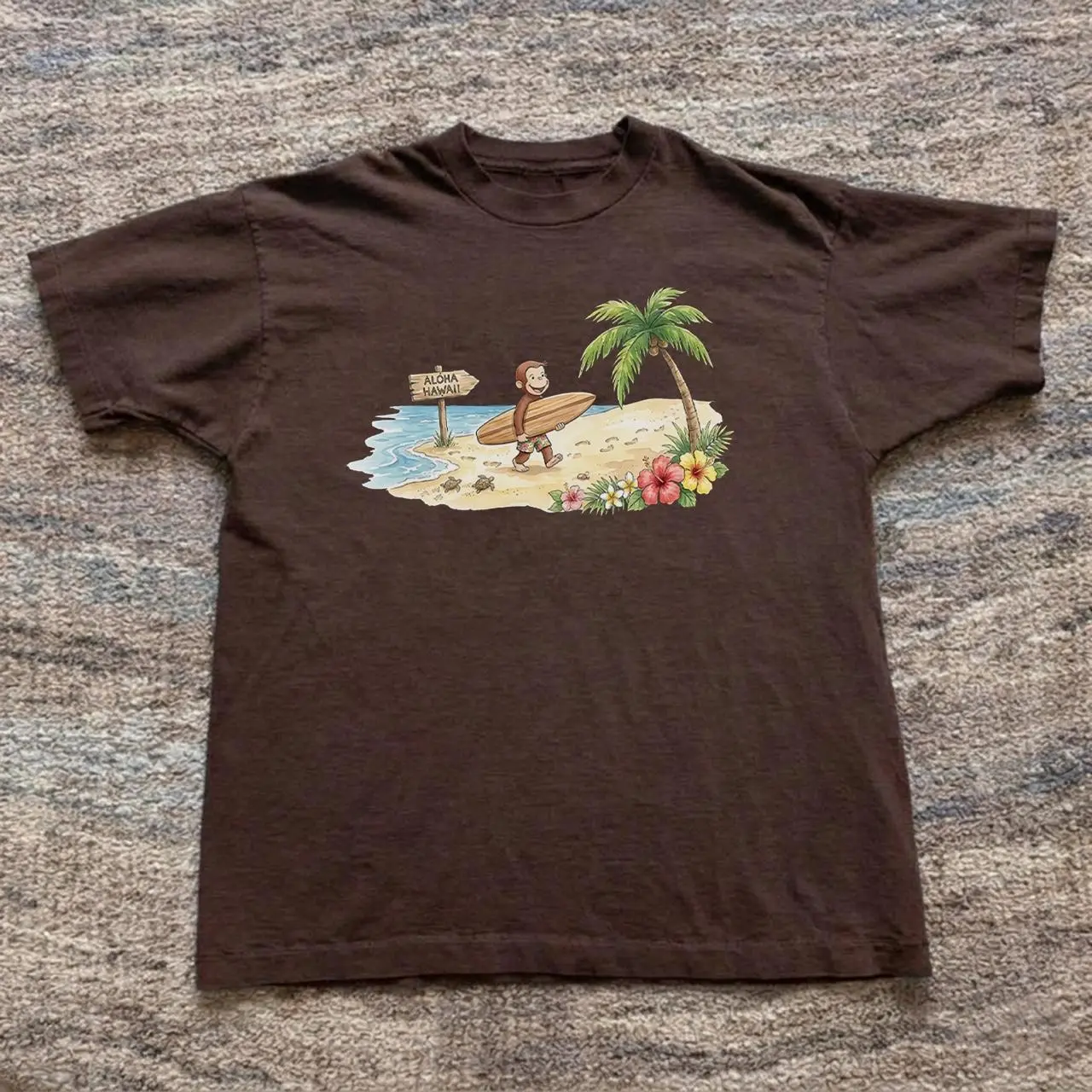 Curious George Aloha Hawaii T-shirt with graphic of a monkey holding a surfboard on a beach with palm trees and flowers