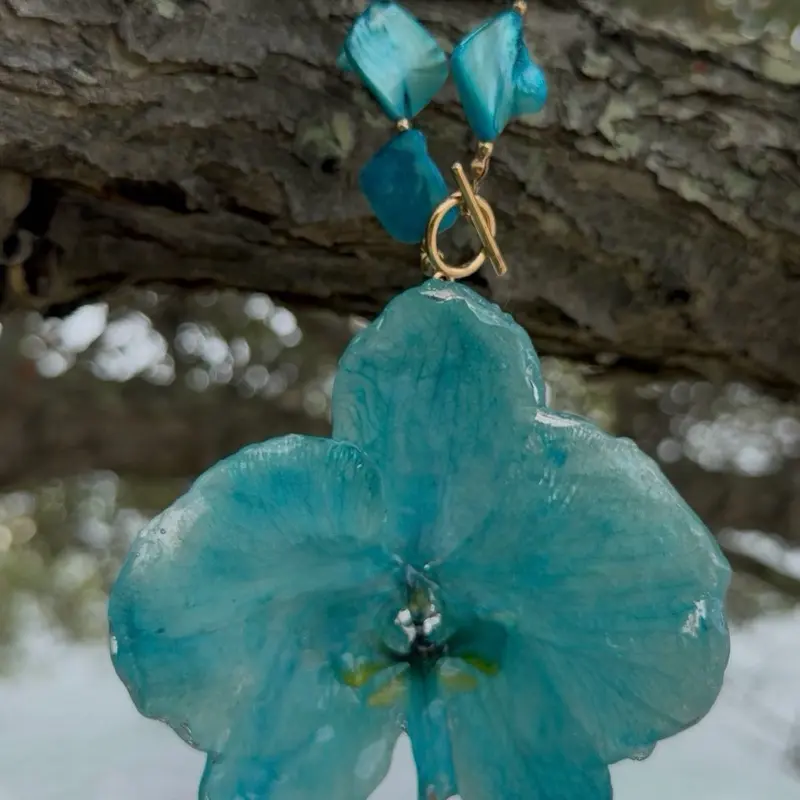 Vibrant turquoise necklace with a large resin-coated orchid flower pendant. The piece has a strand of iridescent, irregularly shaped shell beads in a matching turquoise hue. A small green gemstone charm hangs from the bottom of the orc...